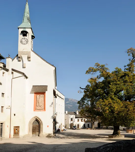Historischer Innenhof mit weißem Kirchturm, großer Baum und Alpenkulisse